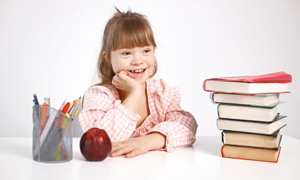 Sunny girl with Down syndrome sitting at desk. Girl studying alone at home. Education for disabled children concept.
